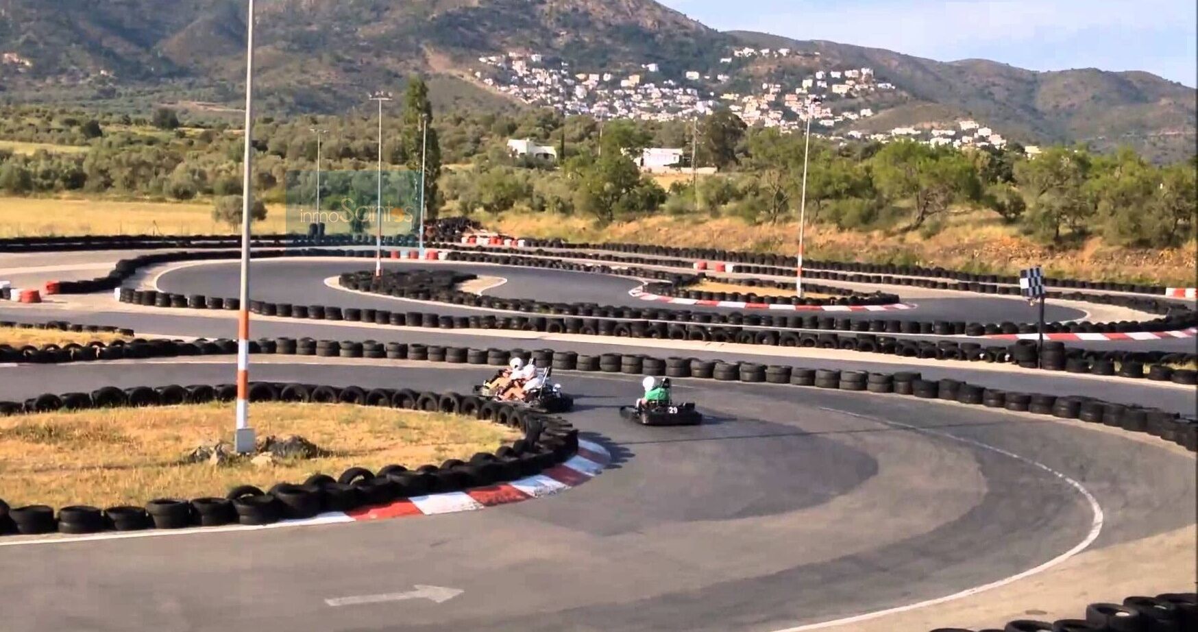 A go-kart track with a single go-kart in motion, surrounded by tire barriers and a mountainous backdrop.