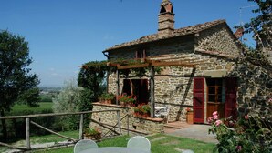 Une maison en pierre avec un patio dans le jardin, des volets rouges et une vue sur la campagne.