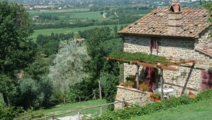 Une maison en pierre avec un toit en tuiles, une pergola en bois ornée de plantes et une vue sur une vallée verdoyante.