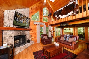 Ein Holzblockhaus-Interieur mit Kamin aus Stein, Ledermöbeln, einem aufgehängten Rindskopf und einem Fernseher, der ein Fußballspiel zeigt.