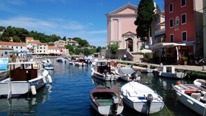 A marina with various boats docked, a prominent pink building with a cross on top, and a tall green tree. - Apartment in Veli Lošinj with Terrace, Air condition, WIFI, Washing machine (63-2) (Veli Lošinj)