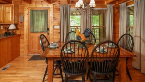 Interior de una cabaña de madera con una mesa de comedor dispuesta para seis personas, rodeada de sillas, un candelabro y una ventana con cortinas.