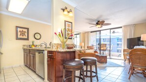 A spacious kitchen with a central island, bar stools, and a dining area with wicker furniture. - Newly Renovated Ocean View Condo 🐳 Stunning Views From The Lanai! 🌅 Kona Reef E-22 (Kailua-Kona)