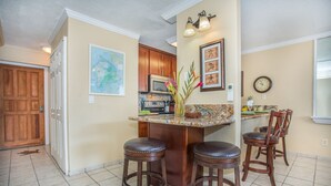 A kitchen with a bar area, wooden cabinets, a microwave, and a clock on the wall. - Newly Renovated Ocean View Condo 🐳 Stunning Views From The Lanai! 🌅 Kona Reef E-22 (Kailua-Kona)