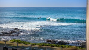 A beach view with waves, a rocky shoreline, and lounge chairs along the edge. - Newly Renovated Ocean View Condo 🐳 Stunning Views From The Lanai! 🌅 Kona Reef E-22 (Kailua-Kona)