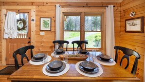 A wooden dining table set with plates, bowls, and glasses, flanked by chairs, in a room with wooden walls and large windows. - Walking Distance to Pool, Putt Putt, Fishing, Ping Pong & more! (Sevierville)