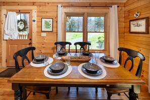 Ein Esszimmer mit einem Holztisch, gedeckten Tellern, Schüsseln und Gläsern, umgeben von Stühlen, an dessen Wänden Holz verkleidet ist und große Fenster vorhanden sind.