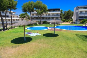 A swimming pool with two umbrellas, a grassy area, and a building with multiple balconies in the background. - Amazing round floor apartment for families - UHC SALOU VILLA 302 (Salou)