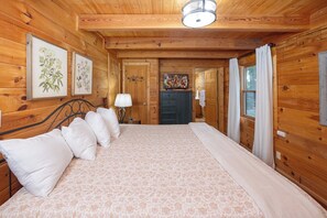 A wooden cabin bedroom with a bed, pillows, a nightstand, a lamp, and framed botanical prints on the wall.