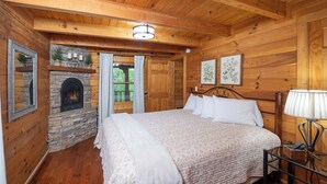 Un dormitorio tipo cabaña de madera con chimenea de piedra, una cama con ropa blanca y una mesita de noche con lámpara.
