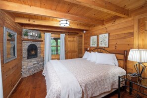 A wooden cabin bedroom with a stone fireplace, a bed with white bedding, and a nightstand with a lamp.
