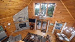 Interior de una cabaña de madera con chimenea de piedra, un comedor y un ventanal con vistas al exterior.