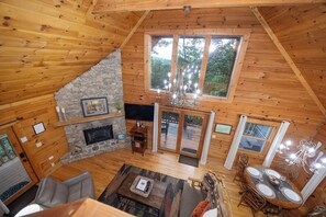 A wooden cabin interior with a stone fireplace, a dining area, and a large window overlooking the outdoors.