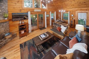 A cozy cabin interior with a stone wall, wooden floors, a fireplace, a television, a sofa, and a dining area.