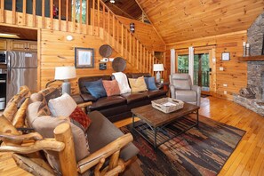 A log cabin interior with a wooden staircase, a living room with a sofa, armchair, and coffee table, and a stone fireplace. - Private w/ Beautiful Views! Hot Tub Looks over Mountains...Beautiful Sunsets!! (Sevierville)