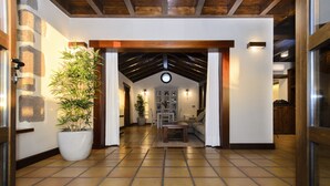 Un pasillo con suelo de baldosas, una planta grande en maceta, techo de madera y vistas a una habitación con sofá y mesa.