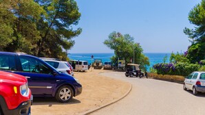 Una strada costiera con auto parcheggiate e un golf cart che conduce a una spiaggia con acque blu cristalline e vista sul mare.