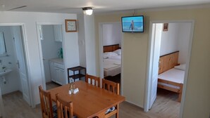 Ein gemütlicher Raum mit Holztisch, zwei Einzelbetten, einem an der Wand befestigten Fernseher, einem kleinen Tisch mit Flasche und Tassen sowie einer Wanduhr.