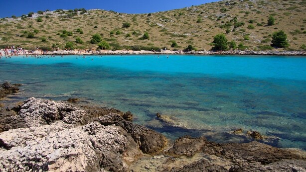 Ein Küstenstrand mit klarem türkisblauem Wasser, felsigem Vordergrund und einer hügeligen Landschaft im Hintergrund.