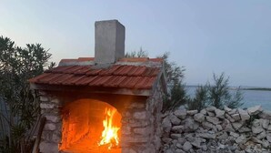 Ein aus Stein gemauerter Außenofen mit Feuer, umgeben von einer Steinmauer und Vegetation.