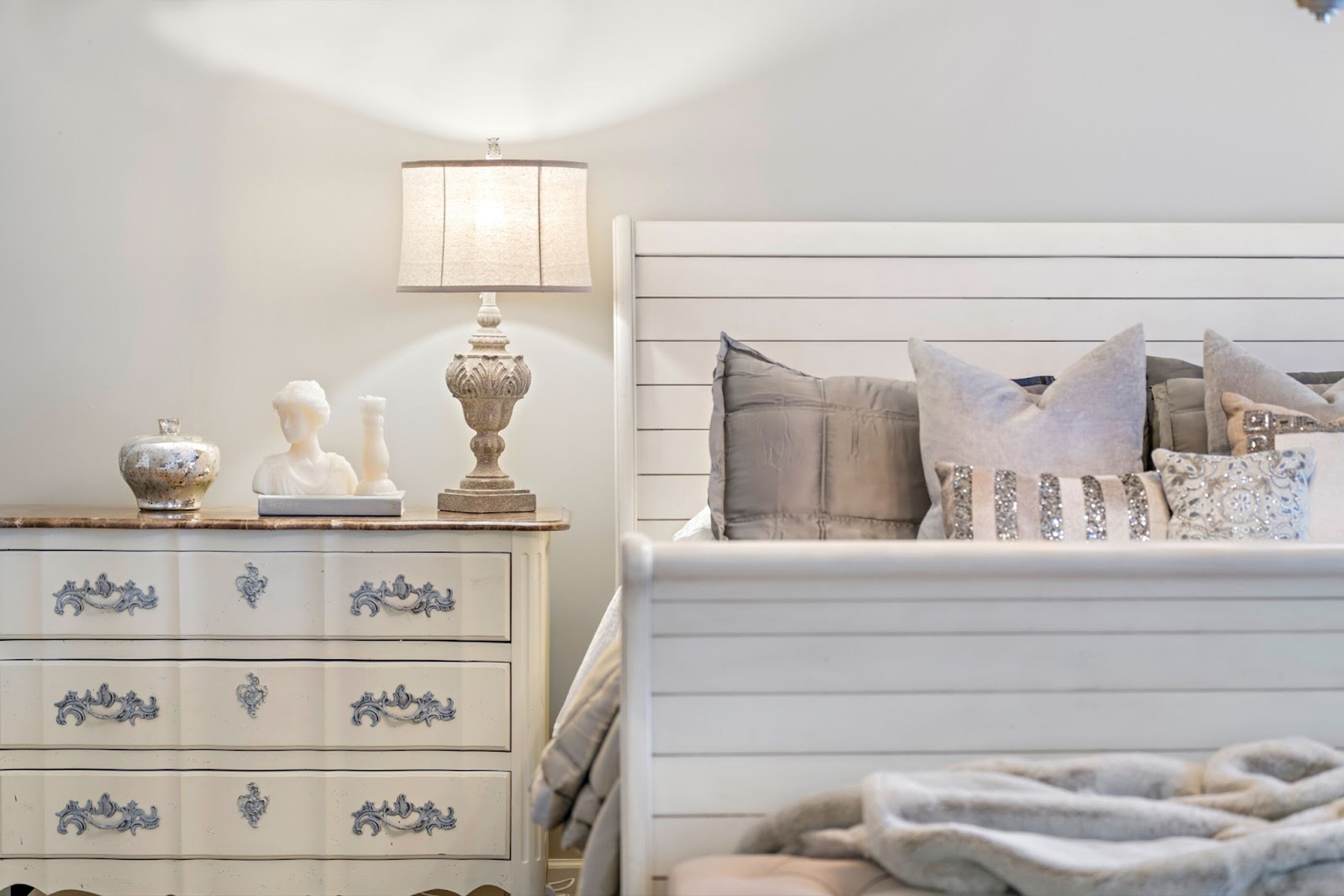 Un dormitorio ordenado con un aparador blanco, una lámpara de noche y una cama con varias almohadas y una manta.