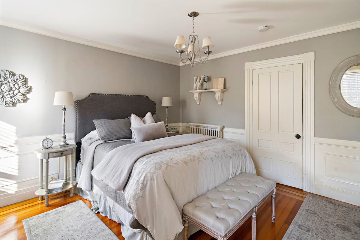 A bedroom with a large bed, bedside tables, a bench, a chandelier, and a door.
