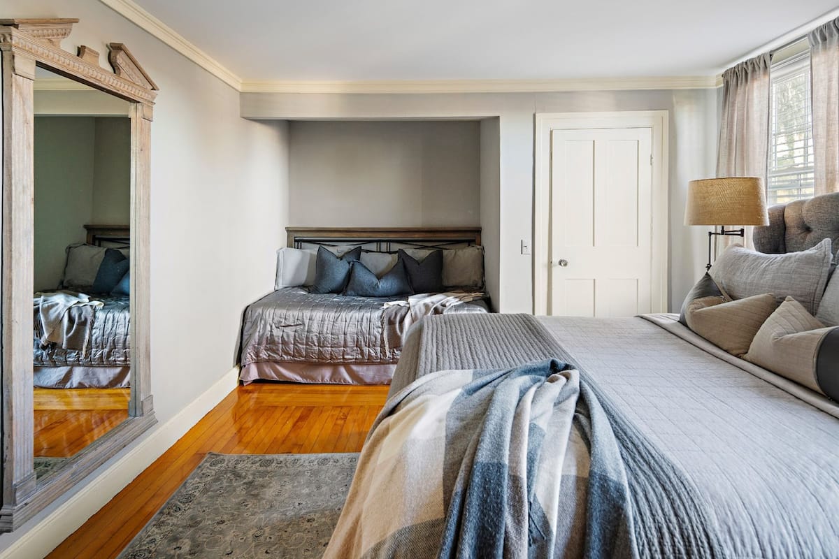 A bedroom with two beds, a large mirror, a bedside lamp, and a view of a hallway.