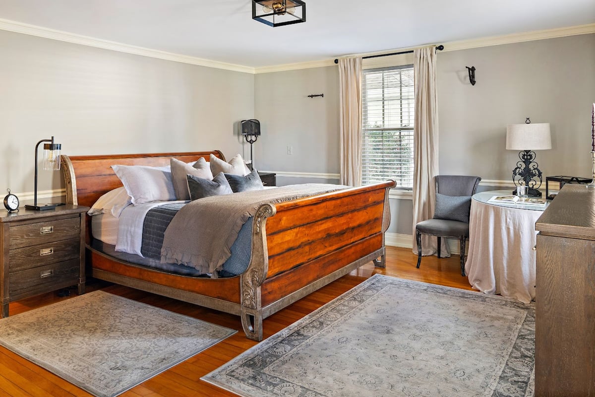 A bedroom with a wooden bed, a nightstand, a chair, a table, and a window with curtains.