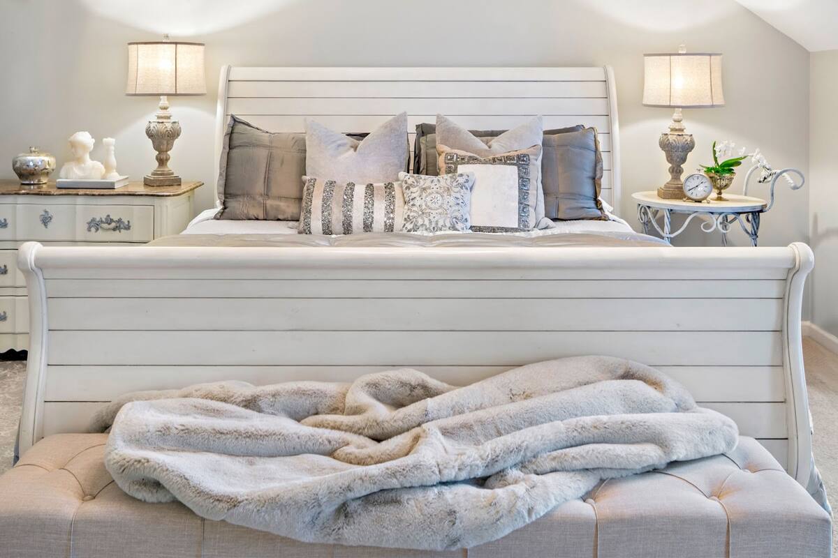 A neatly made bed with a fluffy blanket and multiple pillows, two bedside tables with lamps, and a small side table with a clock and a plant.