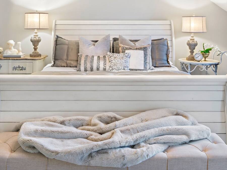 Una cama bien hecha con un edredón esponjoso y varias almohadas, dos mesitas de noche con lámparas y una pequeña mesita con un reloj y una planta.