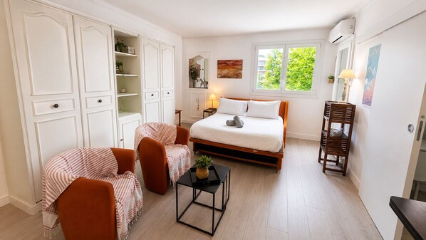 A bedroom with a bed, two chairs, a small table, a mirror, and a window.