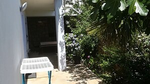 Un étroit sentier extérieur mène à l’entrée d’un bâtiment. On y trouve une petite table et des chaises sur la gauche, le tout étant entouré d’une végétation luxuriante et agrémenté d’un pot de fleurs suspendu.