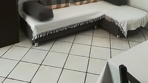 Une petite pièce moderne, dotée d’un canapé-lit, de deux coussins à carreaux, d’une table blanche et d’une fenêtre ornée de rideaux légers.