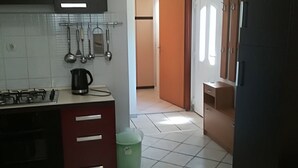 Une cuisine compacte équipée d’une cuisinière à gaz, d’une bouilloire et de divers ustensiles. On y trouve également une poubelle et une petite table avec une chaise.