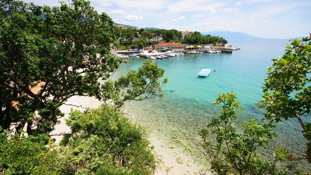 Une vue sur la côte, avec une eau cristalline, une plage de sable et un port de plaisance rempli de bateaux.