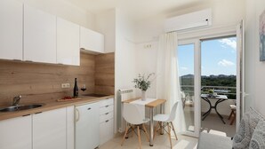 Cocina compacta con armarios blancos, encimera de madera y zona de comedor con mesa y sillas. Se observa un radiador, y la habitación tiene un amplio ventanal corredera que da a un balcón con vistas al paisaje.