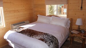 A wooden cabin bedroom with a bed, a nightstand with a lamp, and a window with a view of the outside.