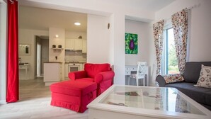 Uma sala de estar com um sofá vermelho, uma mesa de centro de vidro e um sofá cinza com almofadas florais. Cozinha ao fundo com armários brancos e uma cortina vermelha.