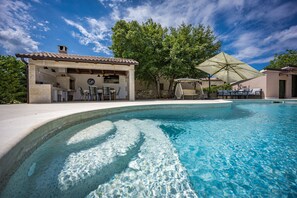 Una piscina con sombrilla, mobiliario exterior y un edificio de piedra con un reloj.
