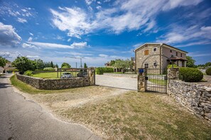 A stone house with a gated driveway, surrounded by greenery and a clear sky.