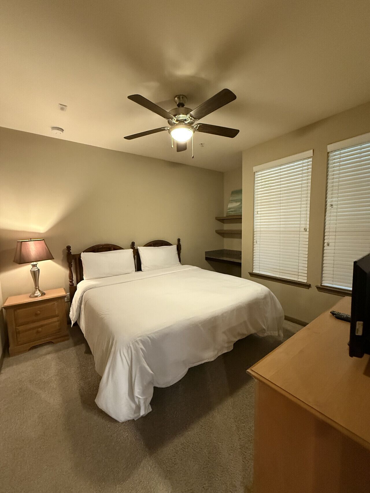 A bedroom with a bed, nightstand, lamp, ceiling fan, and a desk with a TV.