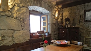 Una habitación con paredes de piedra, una mesa de madera, un jarrón con flores y una ventana con vistas.