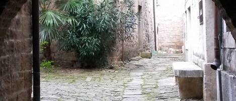 Un pasaje de piedra en arco con un camino adoquinado que lleva a un patio con vegetación y un edificio al fondo.