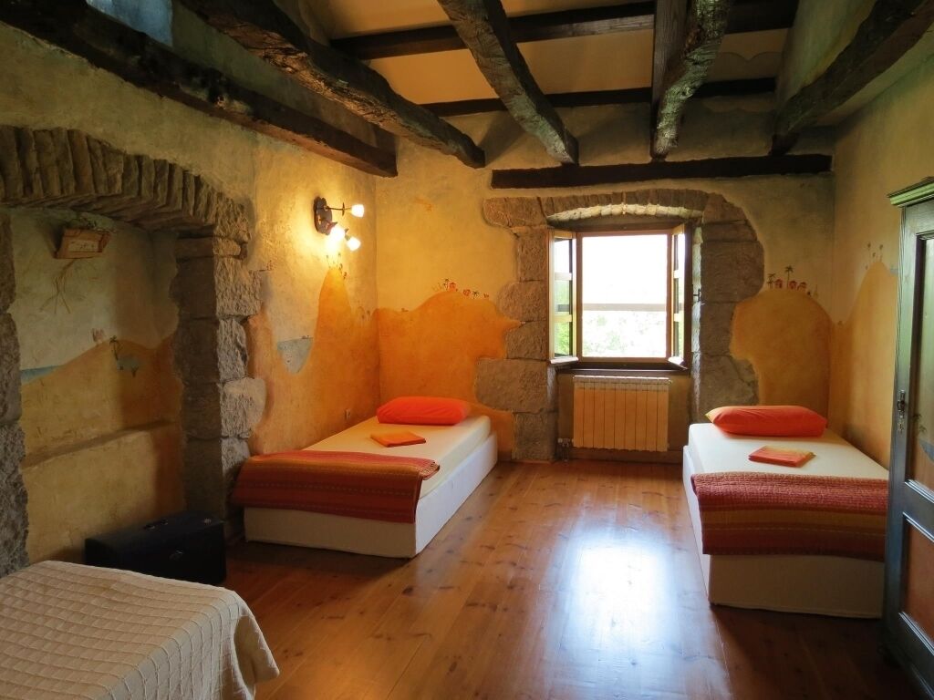 A rustic room with two beds, stone walls, wooden beams, and a window providing natural light.