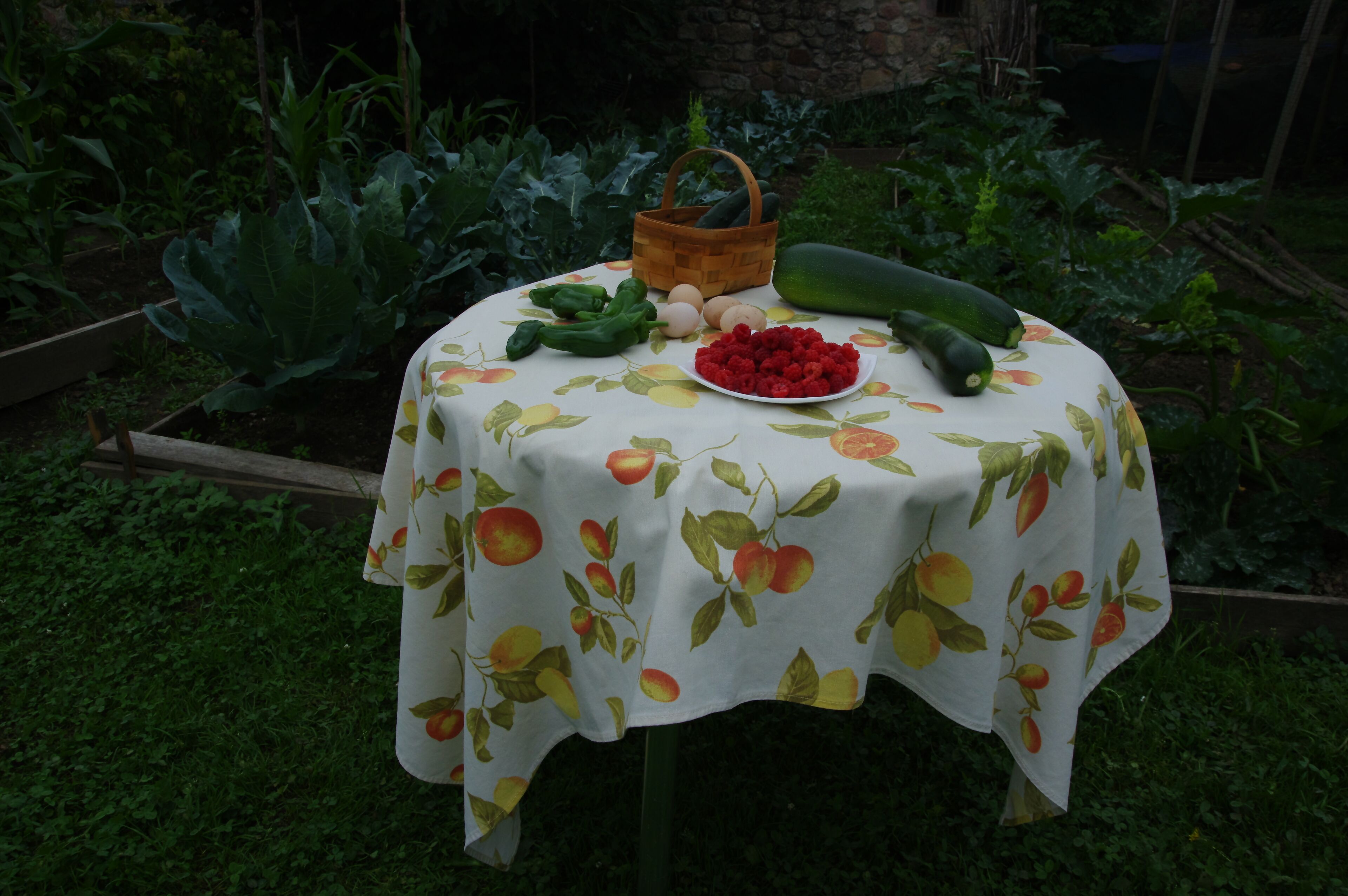 A round table with a tablecloth featuring a fruit pattern, a basket, a plate of raspberries, a cucumber, and some green vegetables.