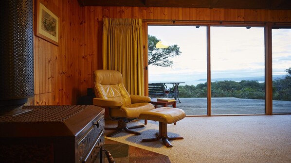 A room with a leather chair, a table with a book and glass, and a view of the outdoors through large glass doors.