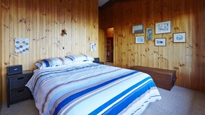 A bedroom with a wooden paneled wall, a bed with a striped bedspread, a bedside table, and a nightstand. - Edith's House (Fairhaven)