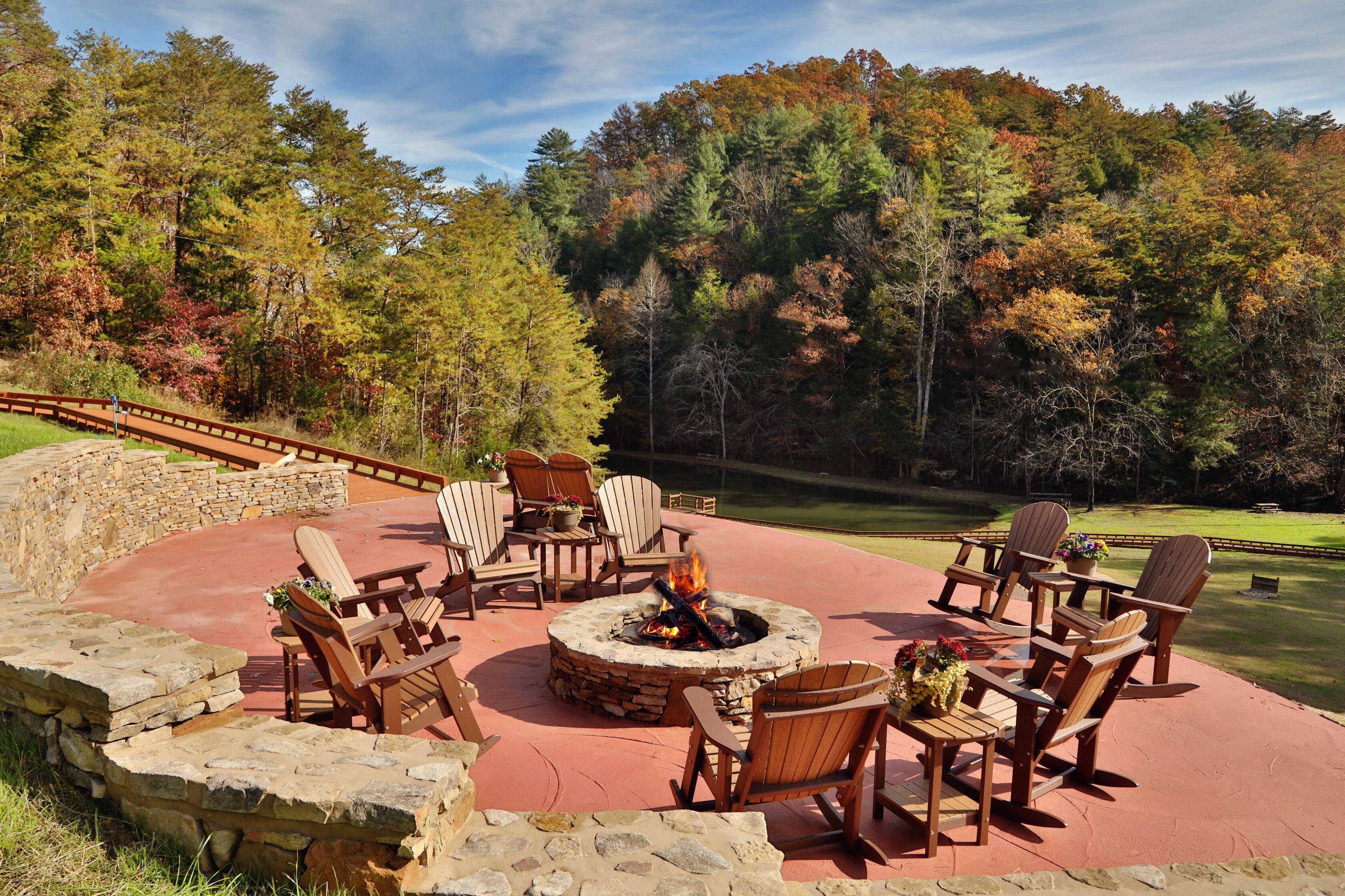Un pozo de fuego de piedra circular rodeado de sillas Adirondack de madera, ubicado en un patio rojo con un muro de piedra y vistas a un campo de golf y un bosque.
