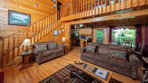 Interior de una cabaña de madera con escalera, dos sofás, una mesa de comedor y una zona de cocina.