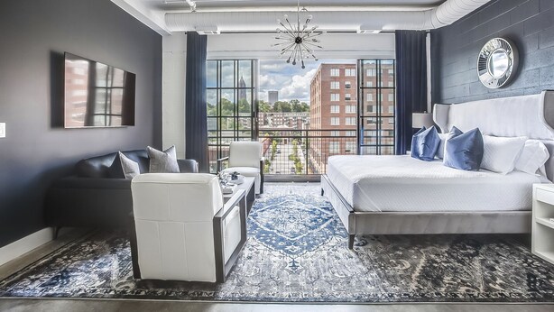 Ein modernes Schlafzimmer mit einem großen Bett, einer Couch, einem Schreibtisch und Blick auf die Stadt durch große Fenster.